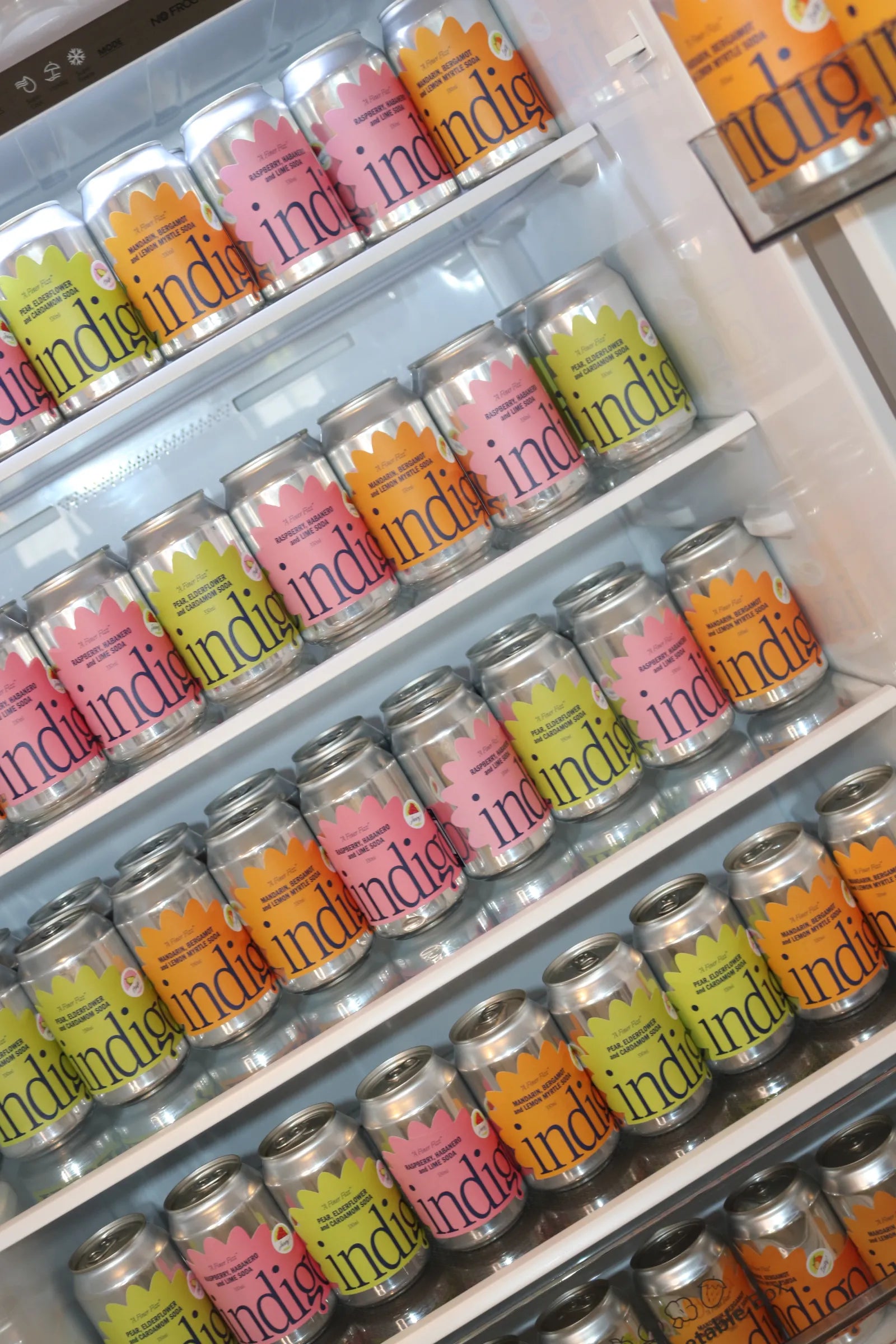 A fridge stacked with indigo botanical soda cans in all three flavours: Mandarin, Bergamot & Lemon Myrtle, Raspberry, Habanero & Lime, and Pear, Elderflower & Cardamom