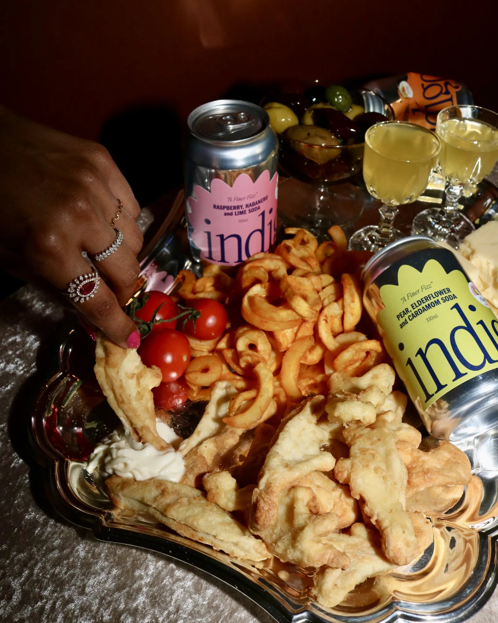 indigo Raspberry, Habanero & Lime and Pear, Elderflower & Cardamom sodas beside a silver platter of fried snacks and tomatoes