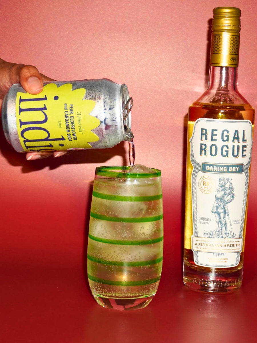 The blanc space – a dry vermouth cocktail. indigo Pear, Elderflower & Cardamom soda poured over ice into a green-striped tumbler, beside a bottle of Regal Rogue Daring Dry vermouth, on a pink and red background