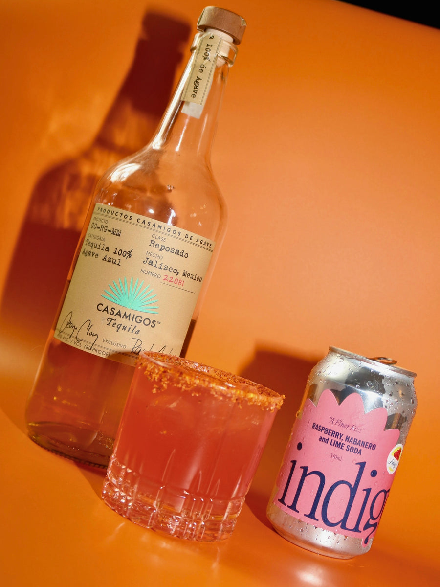 The Second Date – a pink tequila cocktail in a Tajín-rimmed rocks glass, between a bottle of Casamigos tequila and a can of indigo Raspberry, Habanero & Lime soda, on an orange background