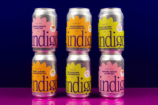 A set of six canned beverages arranged in two rows with three cans in each row. The cans are labeled 'Indigo' with different flavors indicated by colour-coded labels.