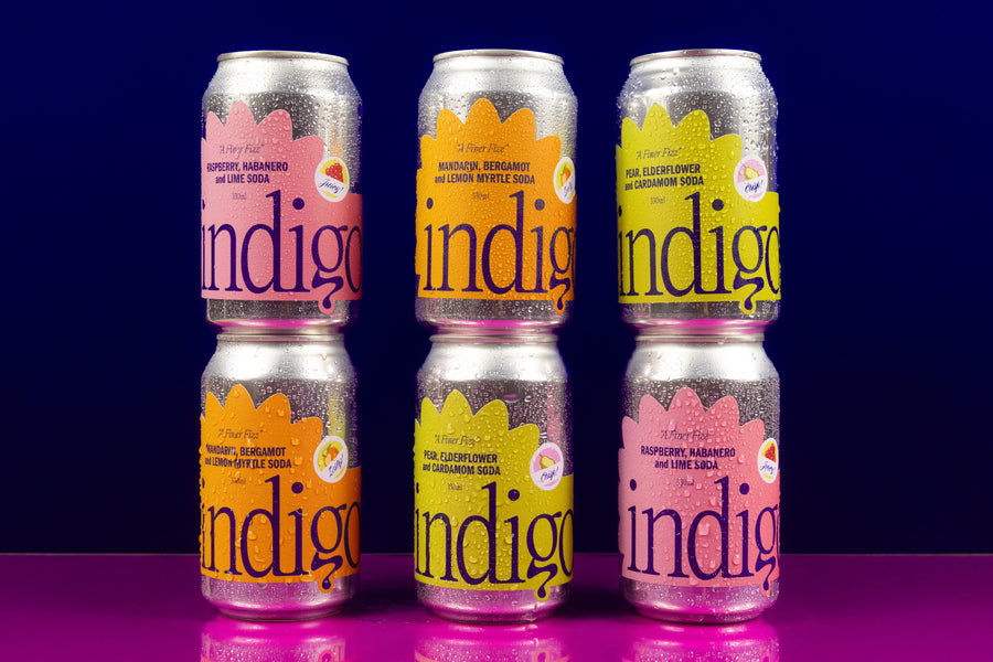A set of six canned beverages arranged in two rows with three cans in each row. The cans are labeled 'Indigo' with different flavors indicated by colour-coded labels.