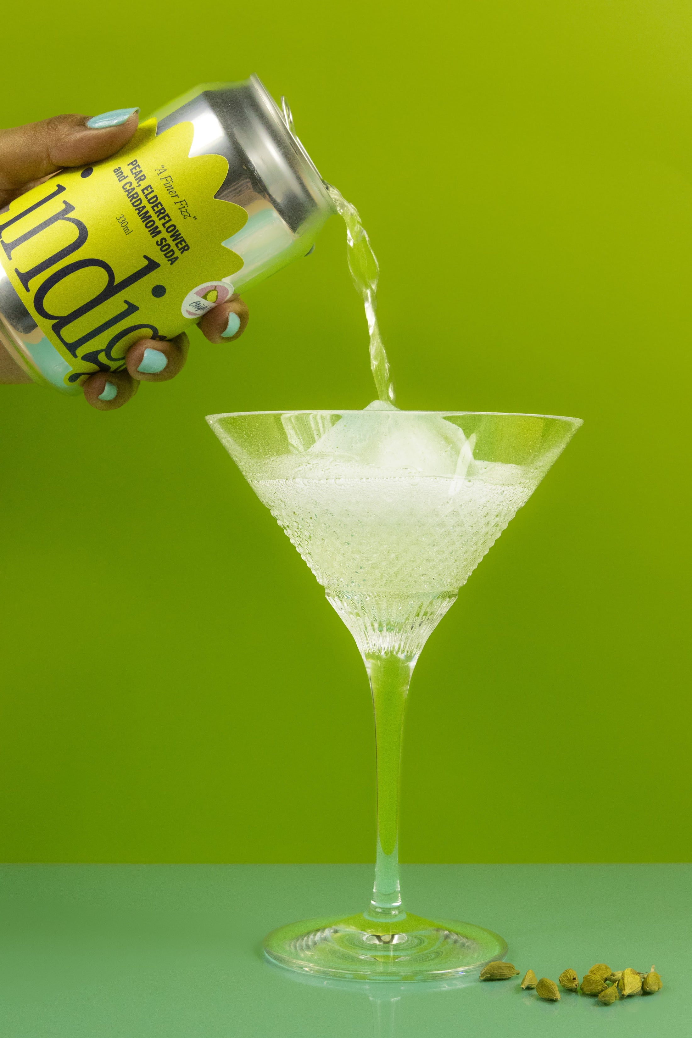 A hand pouring a can of indigo's pear, elderflower and cardamom soda into a tall glass, there are cardamom pods on the side