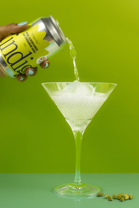 A hand pouring a can of indigo's pear, elderflower and cardamom soda into a tall glass, there are cardamom pods on the side
