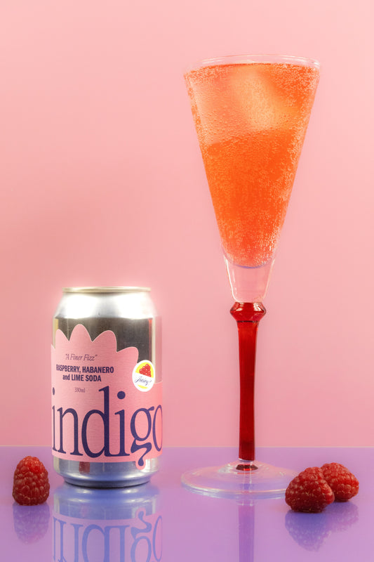 A can of Indigo soda with raspberries in the foreground, beside the can is the soda in a glass with bubbles