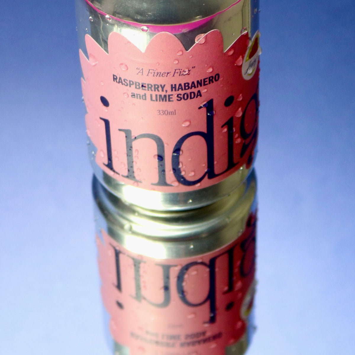 A cold can of indigo Raspberry, Habanero and Lime Soda with water droplets, reflected on a glossy blue surface