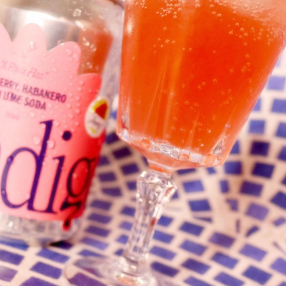 A pink-orange glass of indigo Raspberry, Habanero and Lime Soda, bubbles rising, beside its can on blue and white tiles