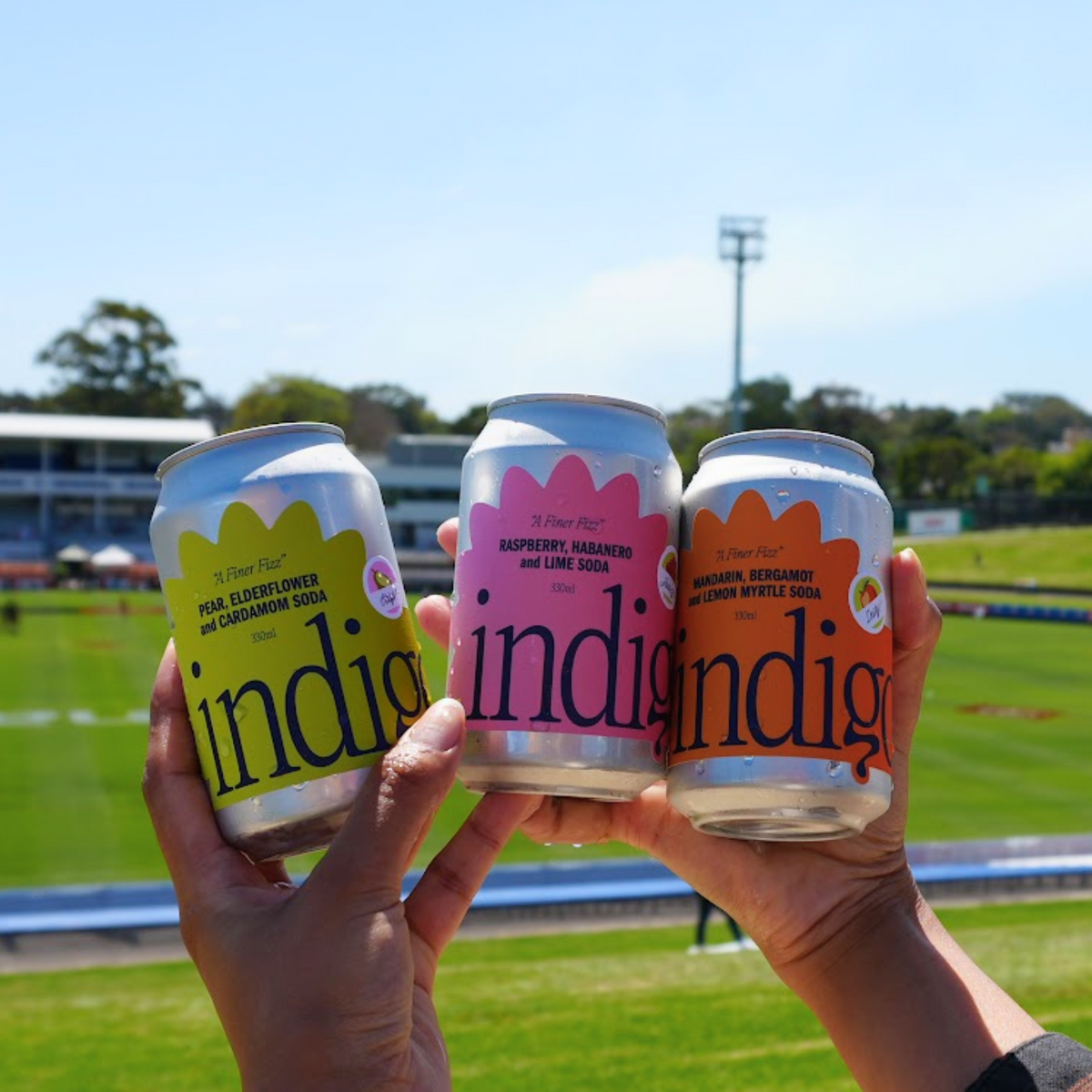 indigo soda Triple Threat mixed pack – Pear Elderflower & Cardamom, Raspberry Habanero & Lime, and Mandarin Bergamot & Lemon Myrtle cans held up at a sunny footy oval