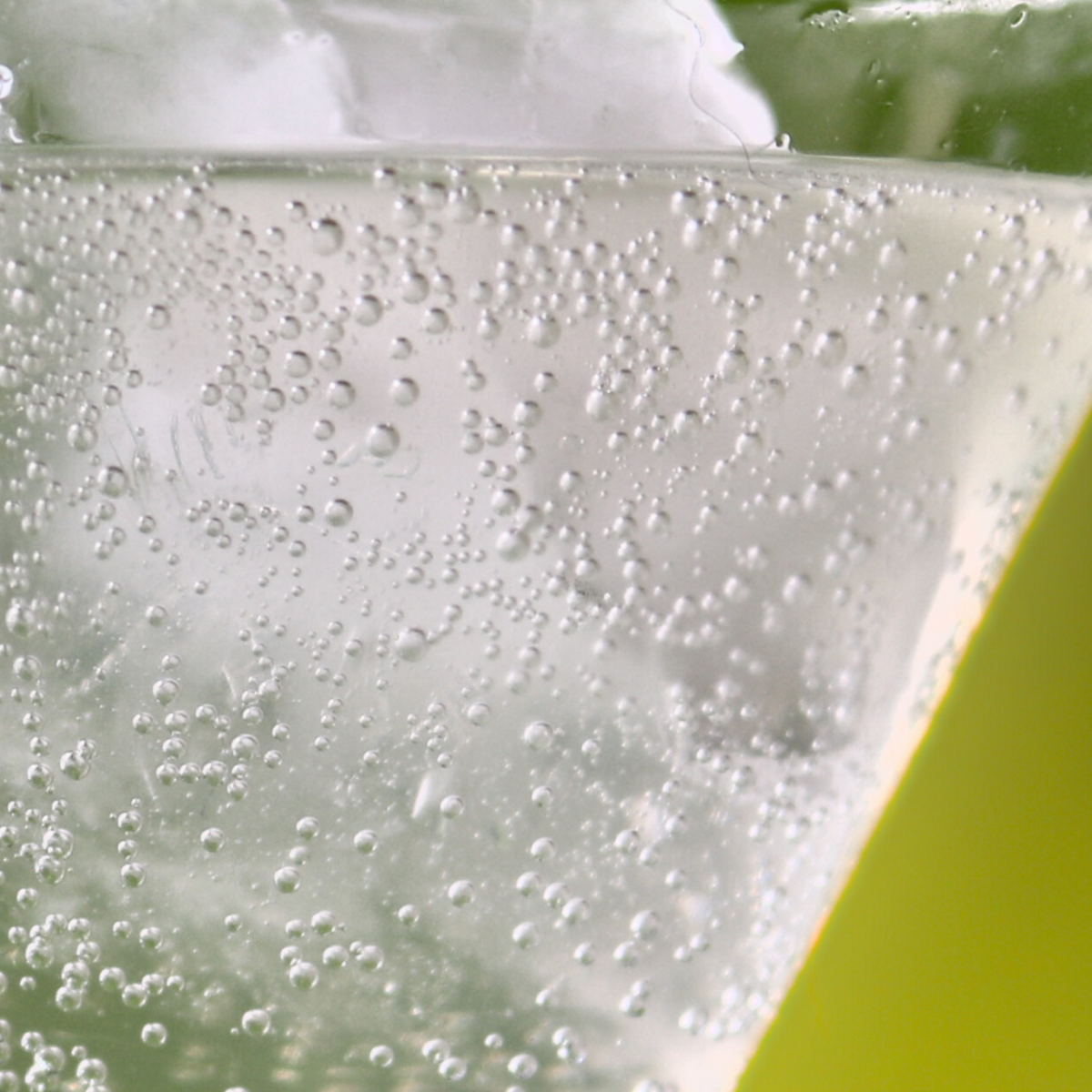 A close-up of fizzing bubbles and ice in a glass of Pear, Elderflower and Cardamom indigo soda against a lime green background